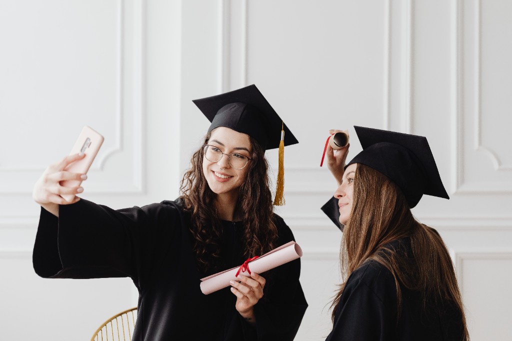 Two graduates in caps and gowns taking a selfie with their diplomas